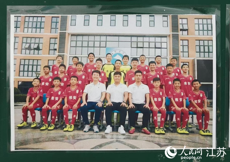 李世恩（上排左四）在淮安工業(yè)園區(qū)實(shí)驗(yàn)學(xué)校05-06組足球隊(duì)的合影張貼在教練室墻上，照片中多名球員正效力于“蘇超”淮安隊(duì)。 人民網(wǎng) 鞠峰攝
